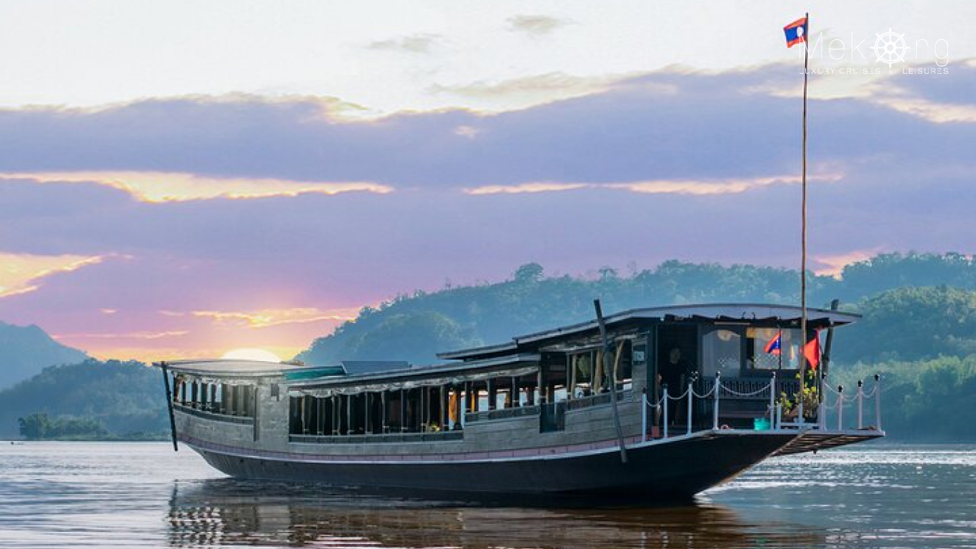 Wooden Mekong Lover Cruise boat flying the Lao flag on calm water at sunset, with forested hills behind