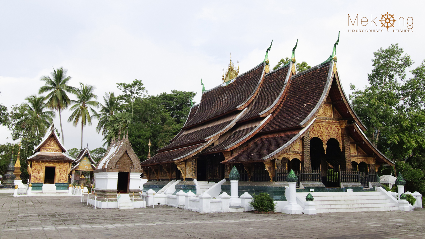 Gilded temple pavilion with a tiered roof, white base and golden naga railings beside tall palms in Luang Prabang