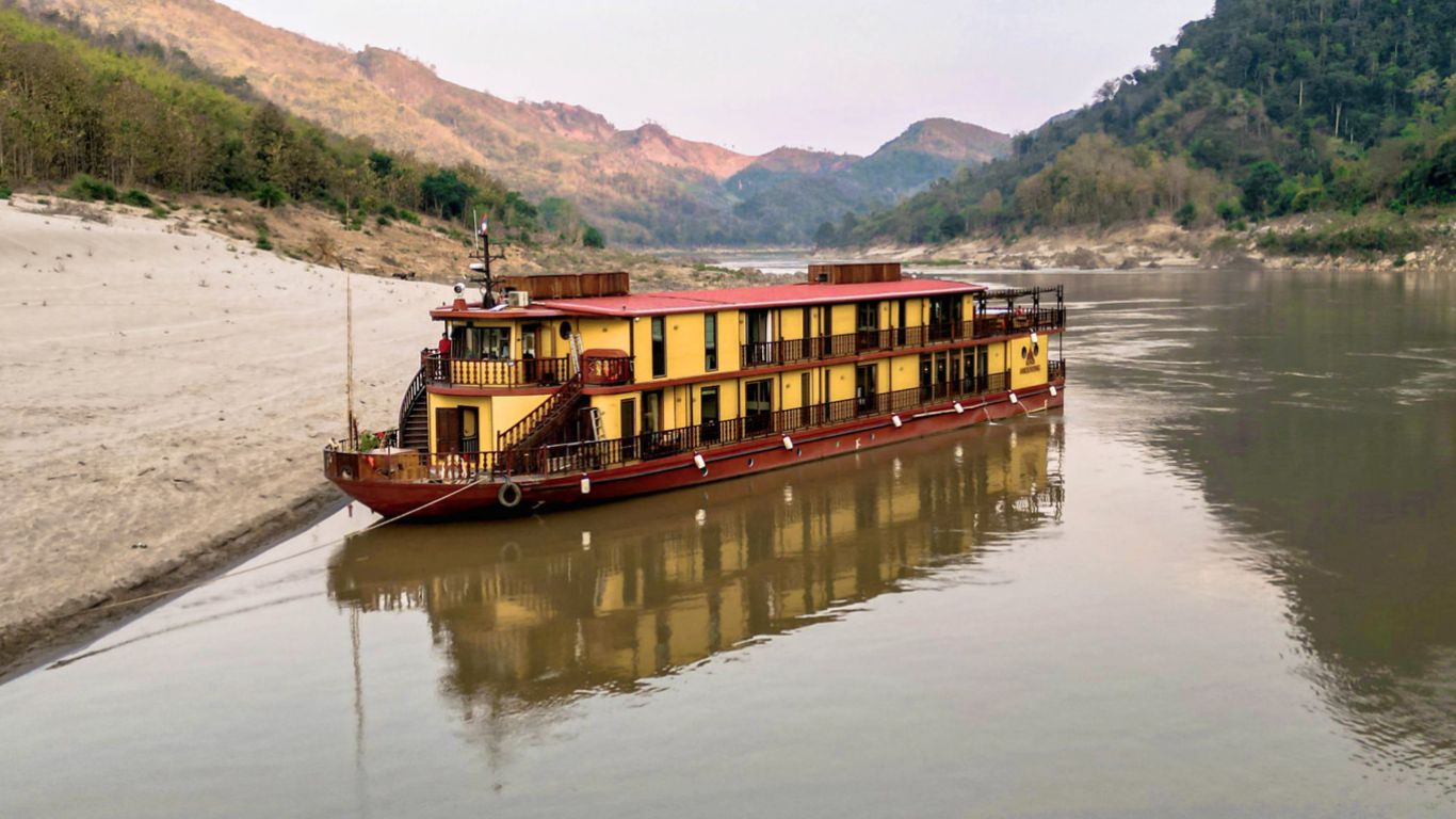 The Heritage Line Anouvong luxury cruise ship docked along a sandy riverbank on the Mekong River, with forested mountains in the background.