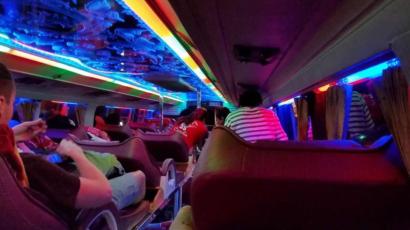Interior of a sleeper bus at night with passengers reclining in bunk style seats under bright multicoloured LED ceiling lights.