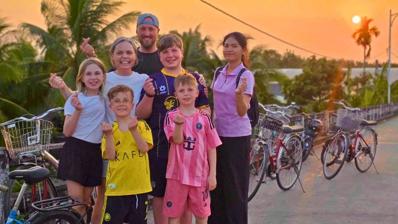 Family with children and local guide posing with bicycles at sunset on a riverside road in the southern Vietnam with orange sky and palm trees