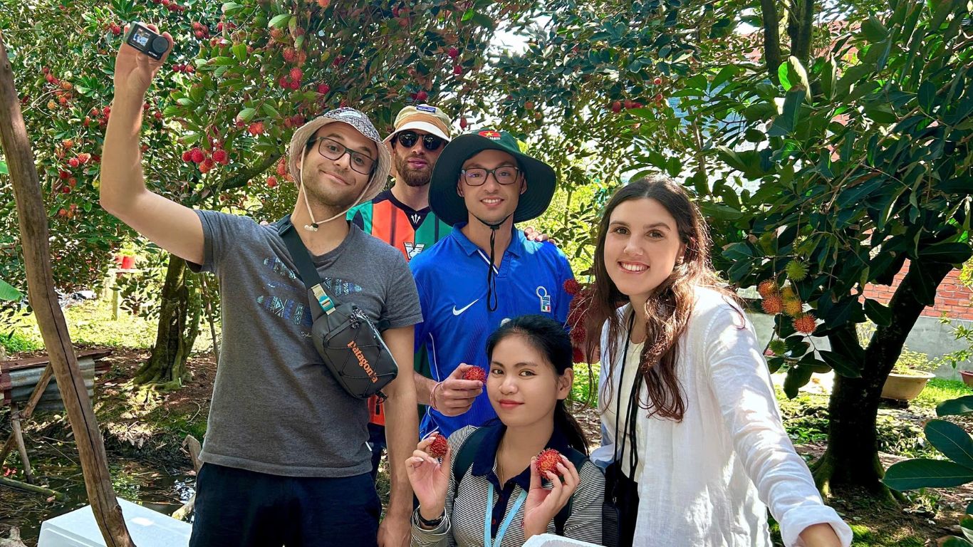 Group of travellers taking a selfie under ripe rambutan trees at a fruit orchard in the Mekong Delta Vietnam with a local guide holding fresh rambutan
