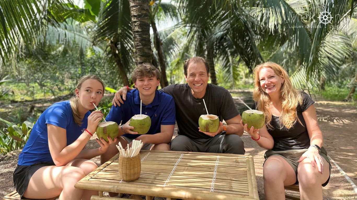 A family of four, including two teenagers, smiling with fresh coconuts at a bamboo table in a coconut grove in the Mekong Delta