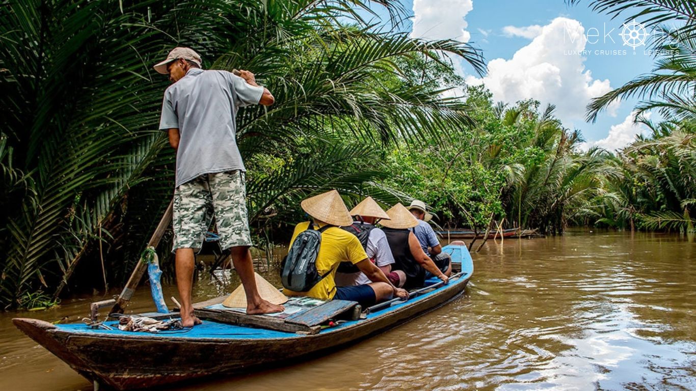 Tourists in conical hats ride a wooden sampan rowed by a local boatman along a narrow canal lined with palms in the Mekong Delta