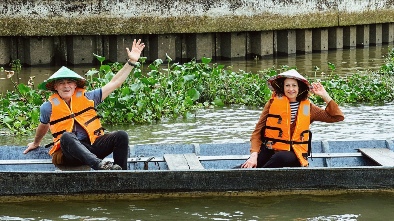 Older couple in orange life jackets and conical hats waving from a small wooden boat beside water hyacinth