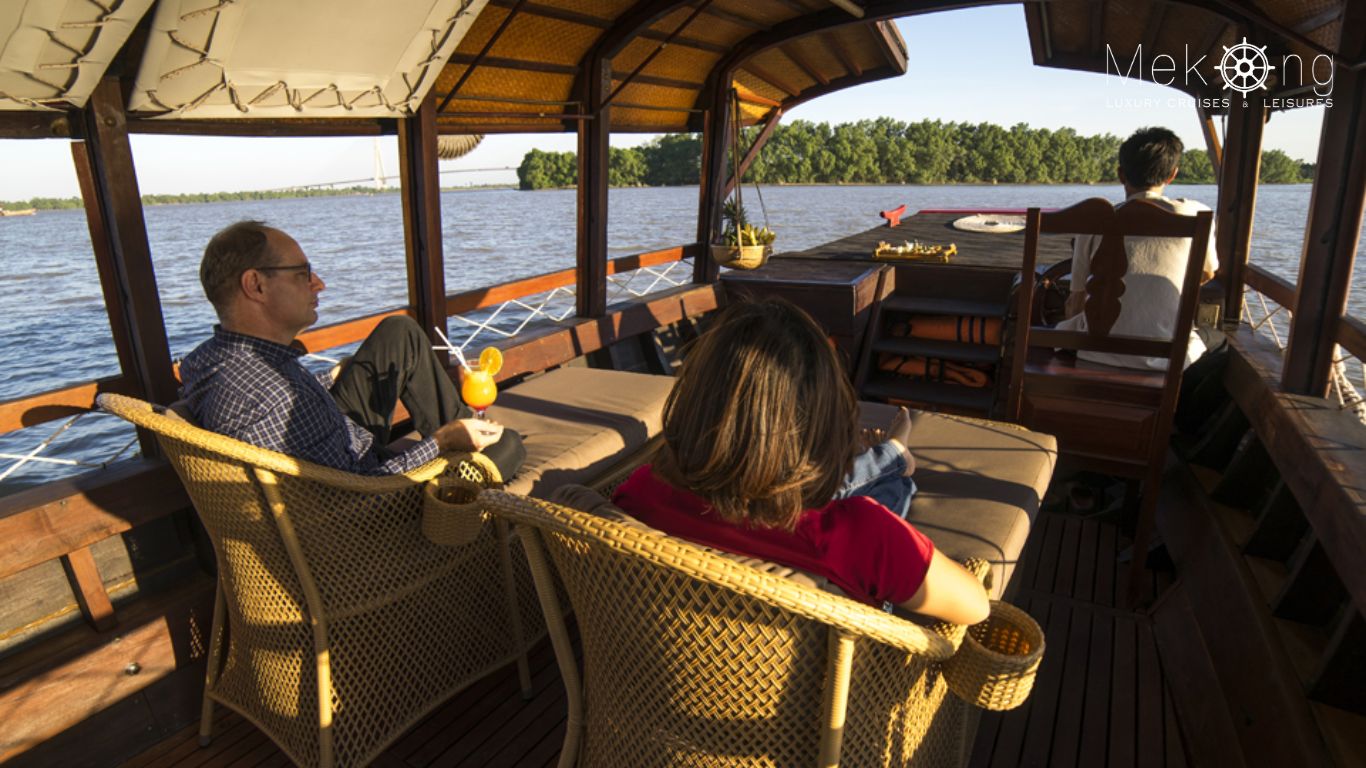 Two travelers relax in wicker chairs on the covered deck of a Song Xanh sampan cruise with a wide river view